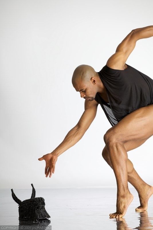 Against a white backdrop, Jeter, a Black dancer, poses on tiptoe, bending & gesturing to a black wicker object on the ground