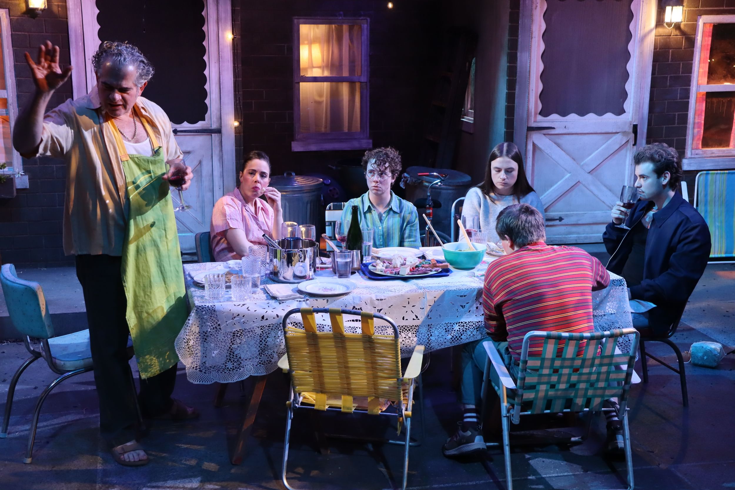 Six people around a festive table with mismatched chairs on a realistic set that looks like a 1970s South Philly backyard.