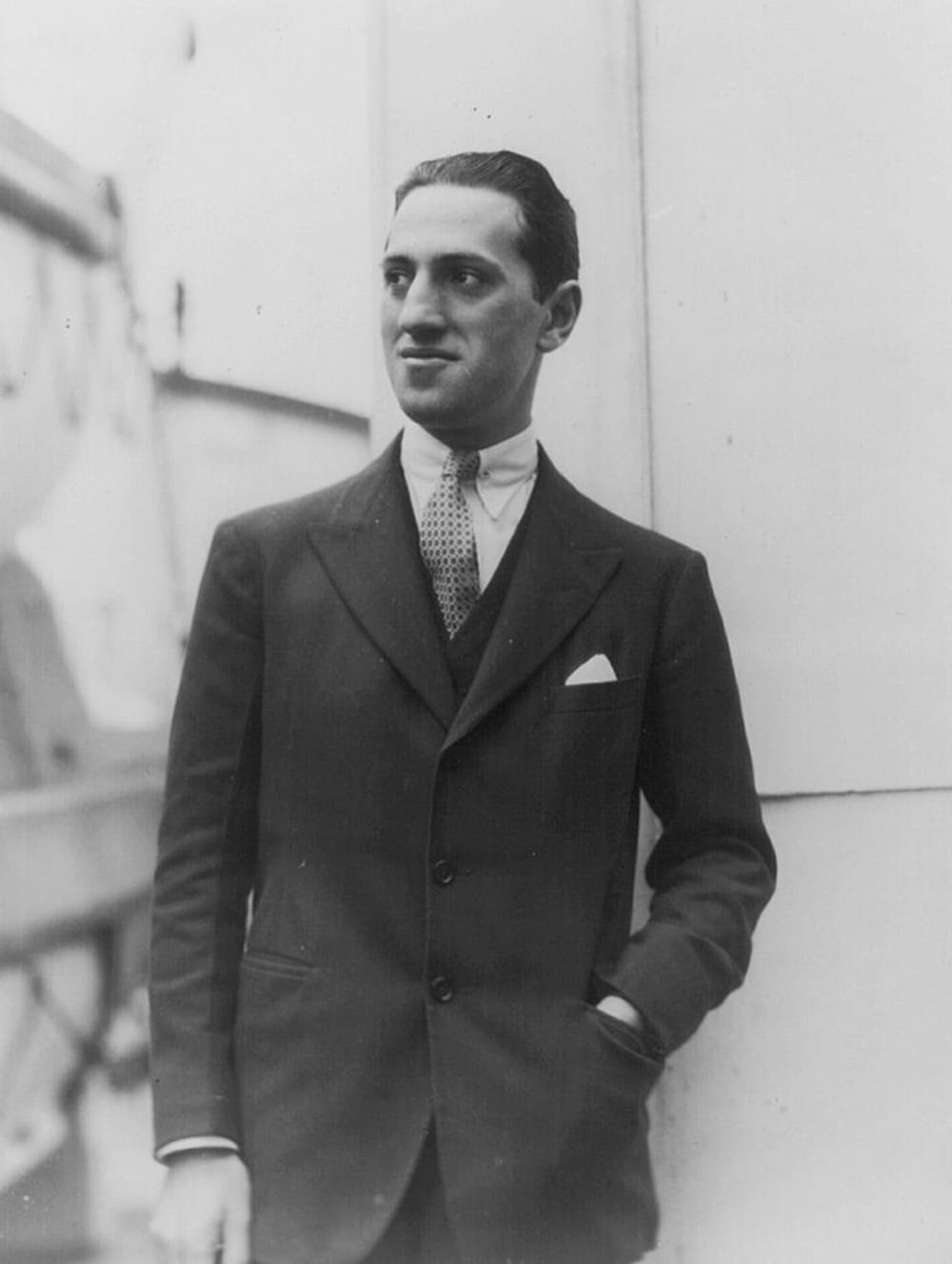 Black and white photo of Gershwin, a Jewish man in a black suit and tie, hand in pants pocket, looking to his left, outside