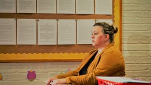 Baldwin-Browns, seen in profile in a mustard-colored top, sits in an elementary classroom chair desk, looking worried.