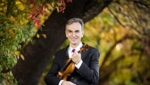 Shaham, 51-year-old white man, stands in front of autumn trees holding his violin and smiling warmly.