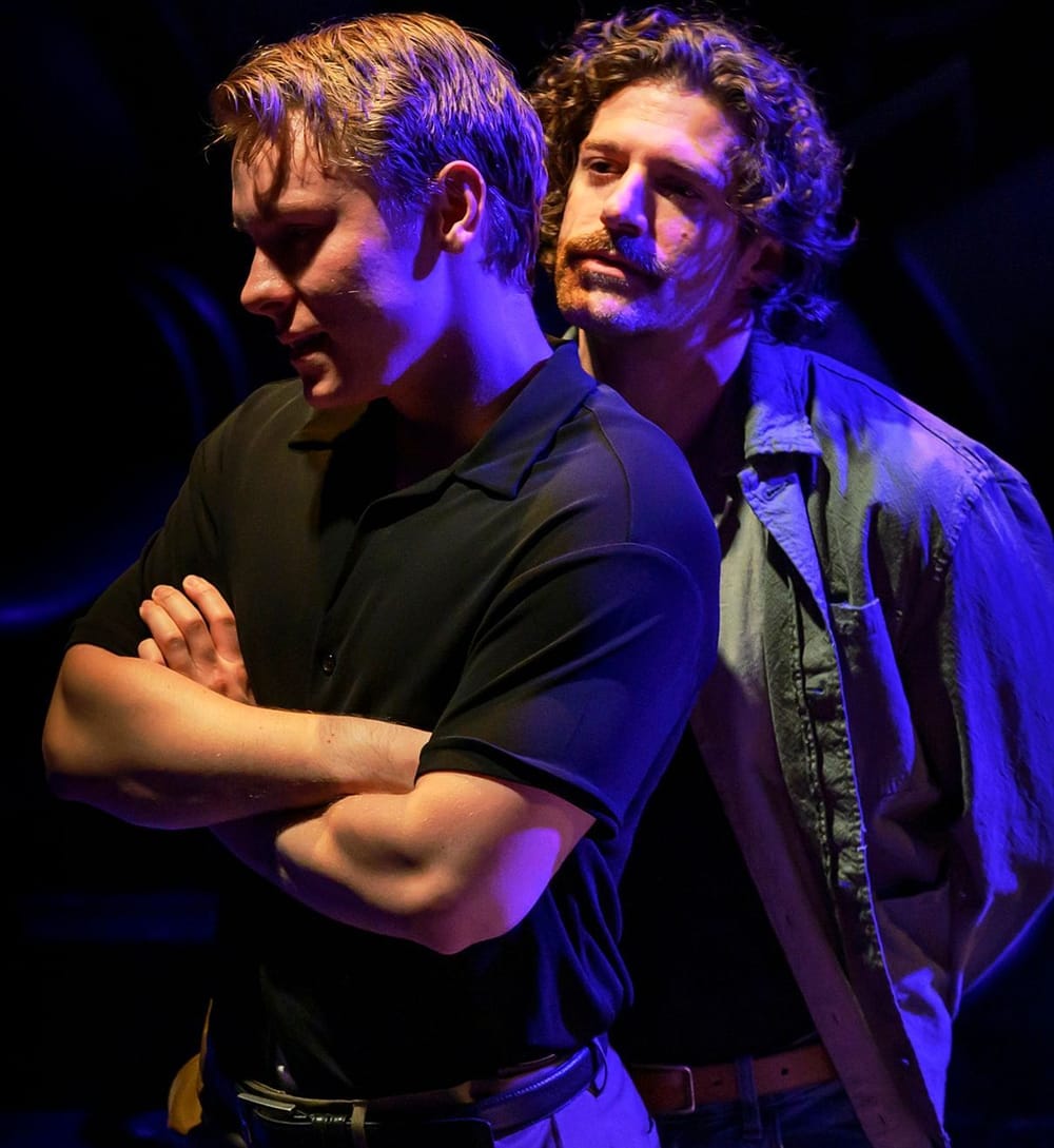 Check, in a black polo, crosses his arms & faces away from mustached Aurelio, who approaches from behind. They're lit in blue