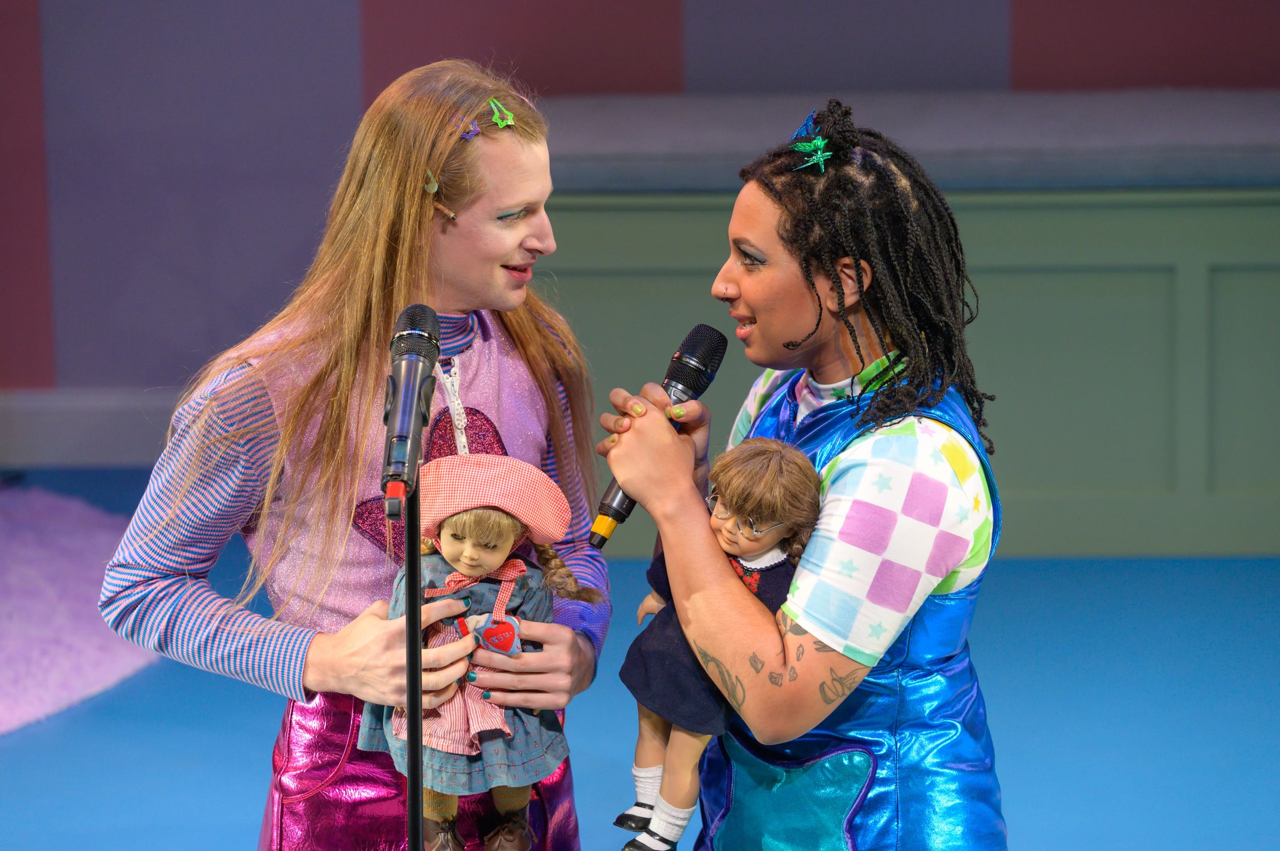 Ressler & Soro face each other smiling, wearing flashy 90s clothes. Soro speaks into a mic. They hold American Girl dolls.