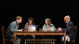 Scene from the musical: the four actors sit smiling around a simply set wooden table, against a black backdrop.