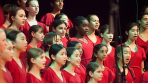 Close-up photo of about two dozen members of the girls’ choir, in matching red dresses, singing together.