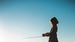 Seen in profile, a little girl in a play dress and bobbed hair stands on the shore of a lake using a kid-sized fishing rod.