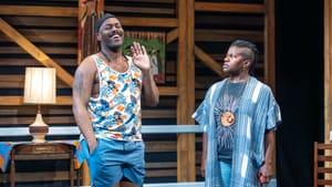 Wilfred, in blue shorts and floral tank, laughs while Kidwell, in jeans and a blue robe, looks on angrily.