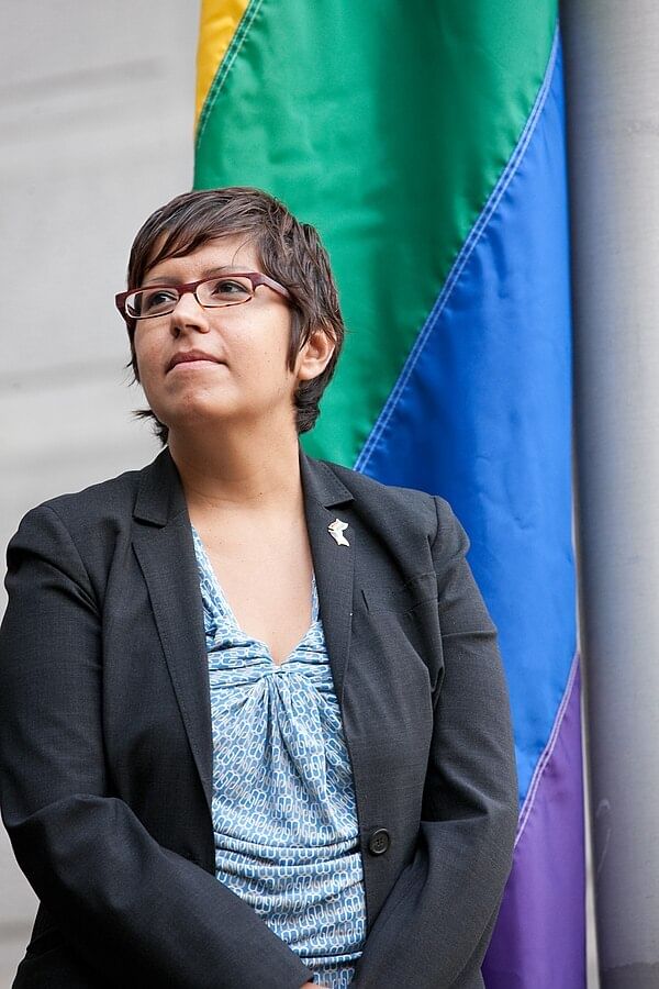 Closeup on Casarez, a Latine woman with glasses, standing solemnly in a dark blazer next to a pride flag.