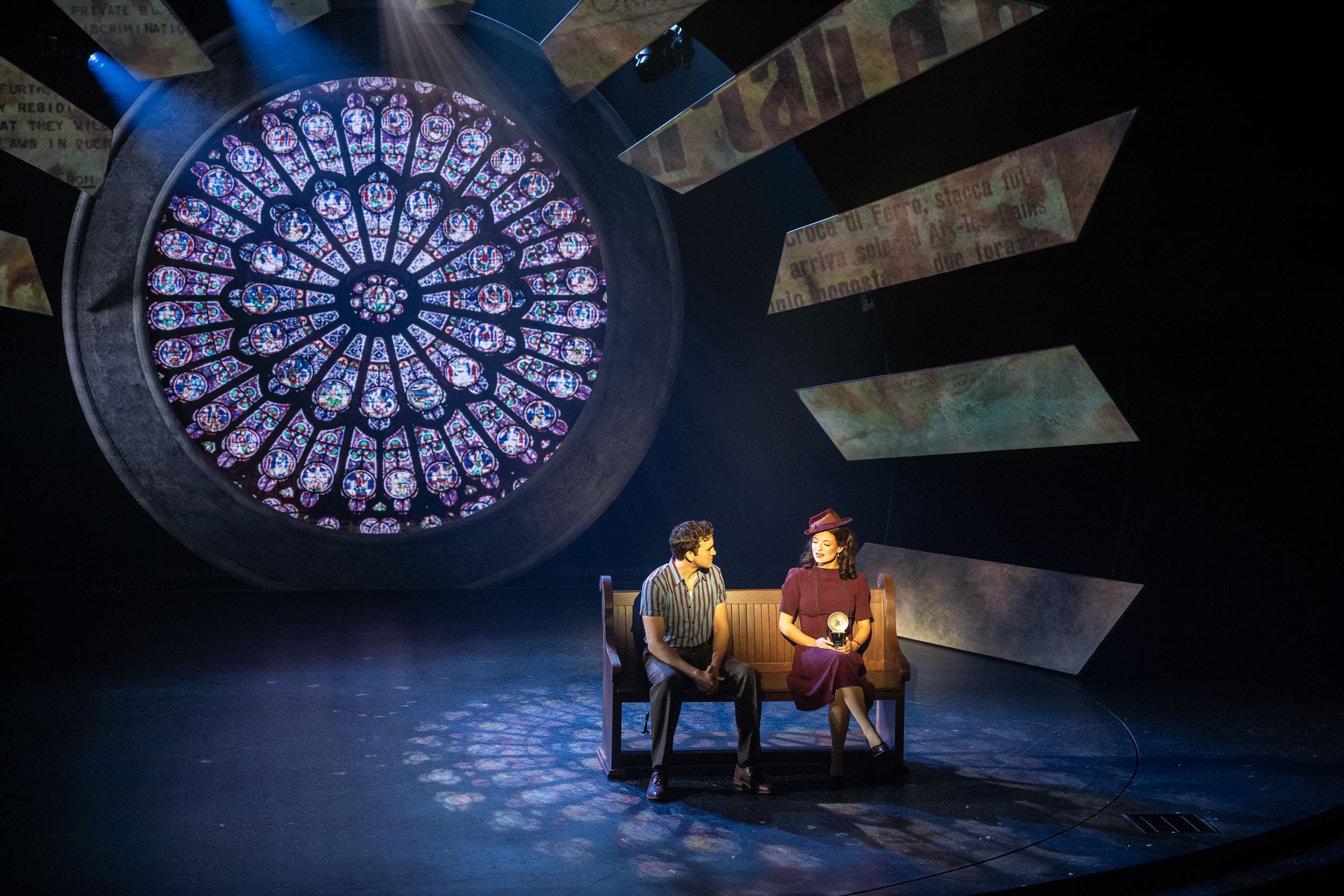 Amira & Prestage, in 1940s clothes, sit together on a bench in the light of large round cathedral-style stained glass window.