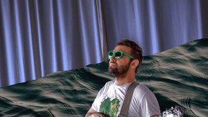 Against a fanciful watery background, Haro poses regally with his guitar on a stool, wearing green-framed sunglasses.