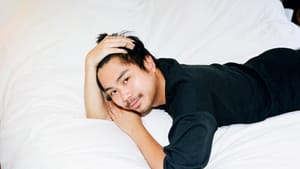 Lee, an Asian man, lies on a white bed, looking up and smiling. He wears a black shirt.