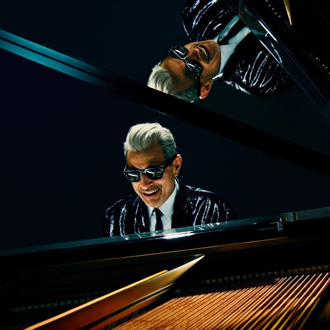 Closeup of Goldblum, grinning in dark glasses and a black sequined blazer, playing a shiny grand piano.