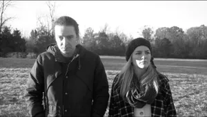 B&W film still. Two people stand next to each other outdoors in daylight, wearing wintry jackets.