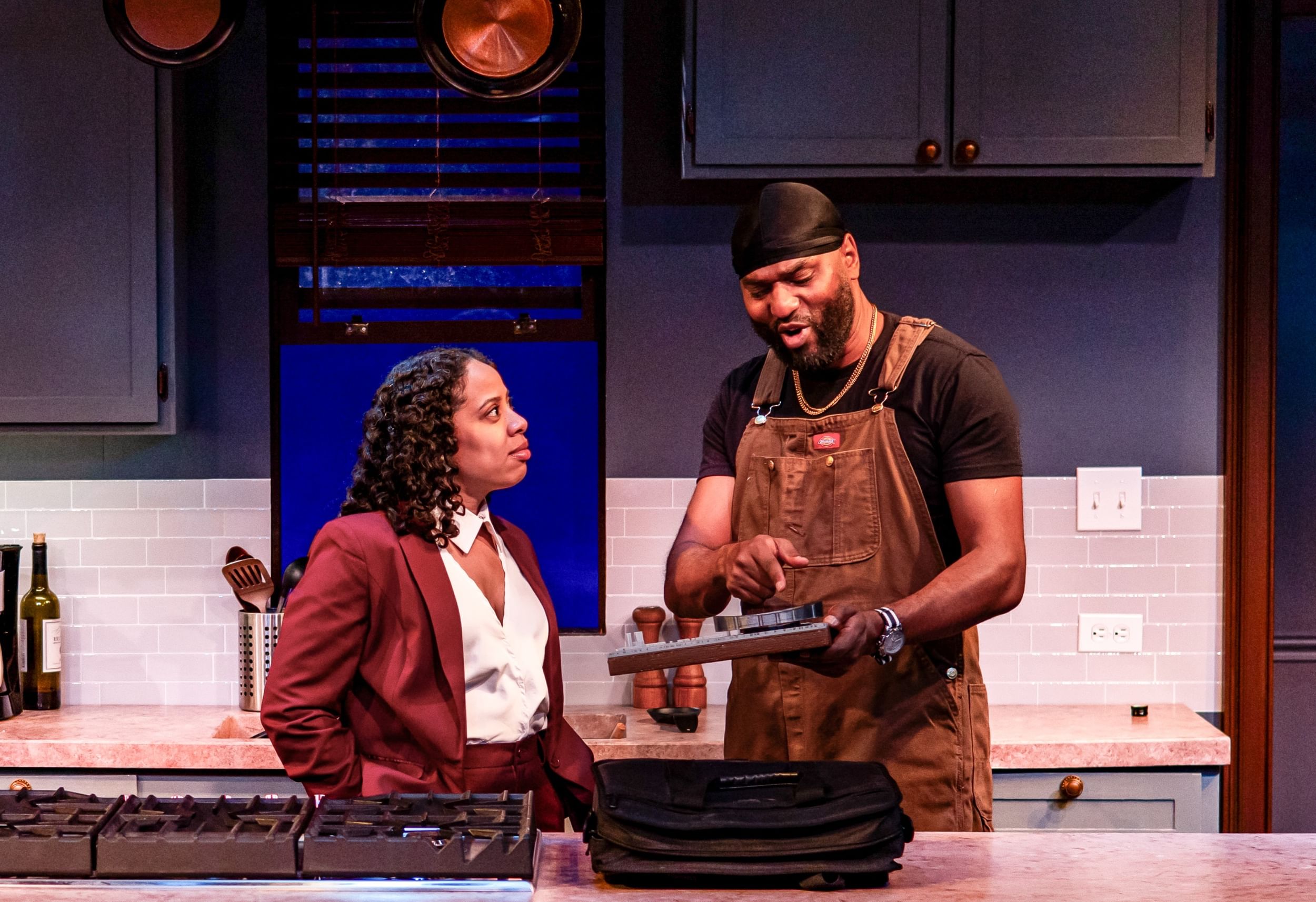 In a modern kitchen, the actors interact over a handheld model of a stadium in a city.