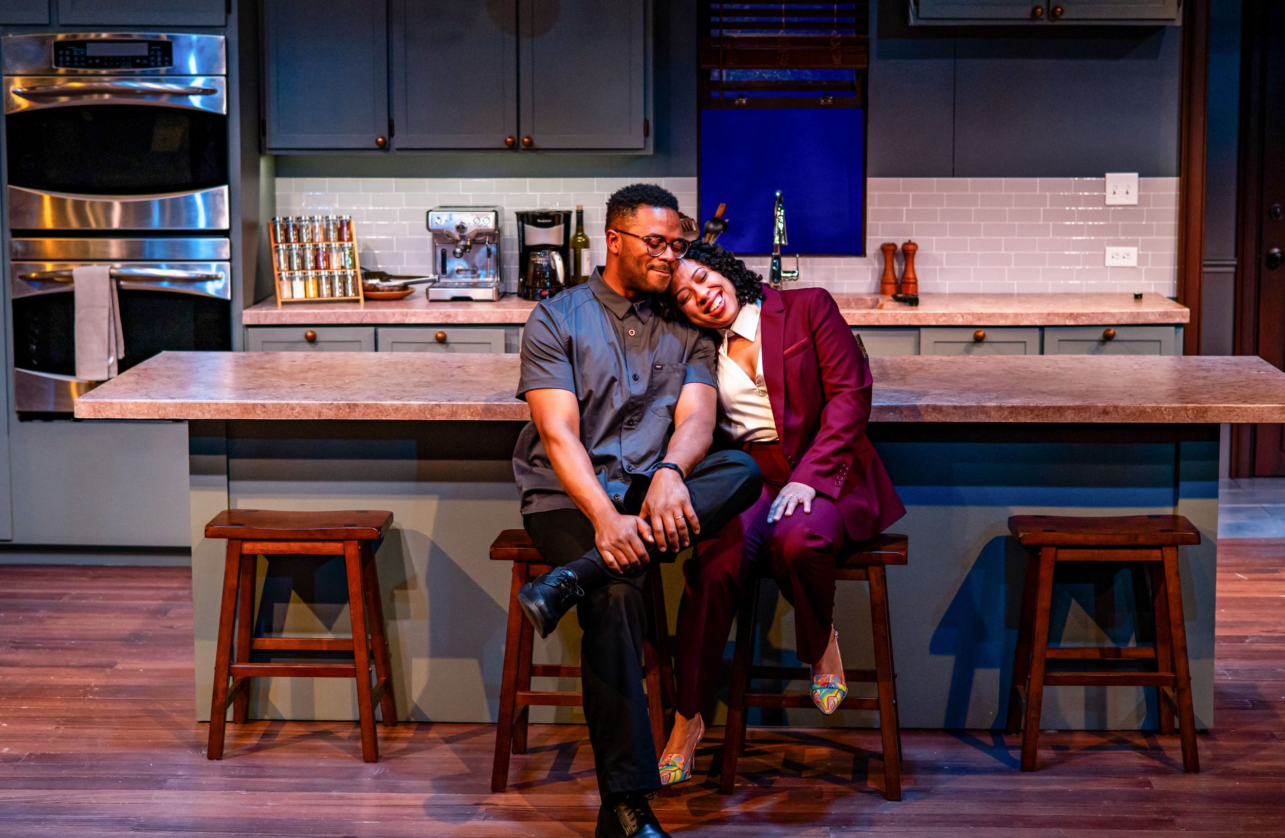 In a spotless, affluent modern kitchen with a large island, the actors cuddle at center, smiling, on wooden stools.