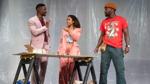 At a construction site, Athie, in a light mauve suit, talks to Watson, in a peach suit. Davis, dressed for work, looks on.