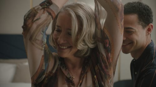 Emma Thompson, with gray hear hair & sheer patterned shirt, lifts arms over her head next to Daryl McCormack. They’re smiling