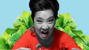 A woman of Asian descent appears to yell, looking at cam, in b&w except for her red floral shirt and plant leaves behind her