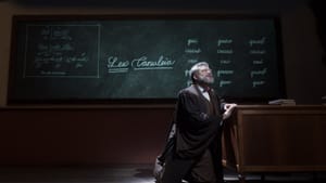 Granner in a judicial robe, on his knees at a desk, soft light cast on him. A blackboard behind him has Latin written on it.