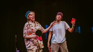 The two actors, dressed in baggy men’s clothes, leer while holding ping pong paddles and red solo cups.