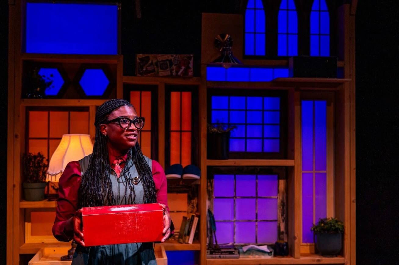 Robinson, a Black woman with glasses and long braids, holds a red box on a stage with fanciful blue and red-lit windows.