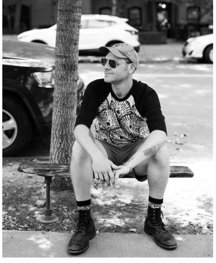 B&W portrait, Labold in casualwear sits on a small bench outside, during daylight, wearing sunglasses looking to his right