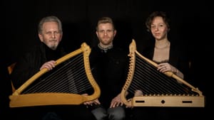 In a mirrored pose, Baby & Črnčič sit holding Medieval harps with leiken between them. They have serious but friendly faces