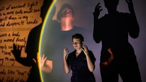 Johnson, in a dark jumpsuit, makes a grieving gesture while their image and their shadows project large behind them.