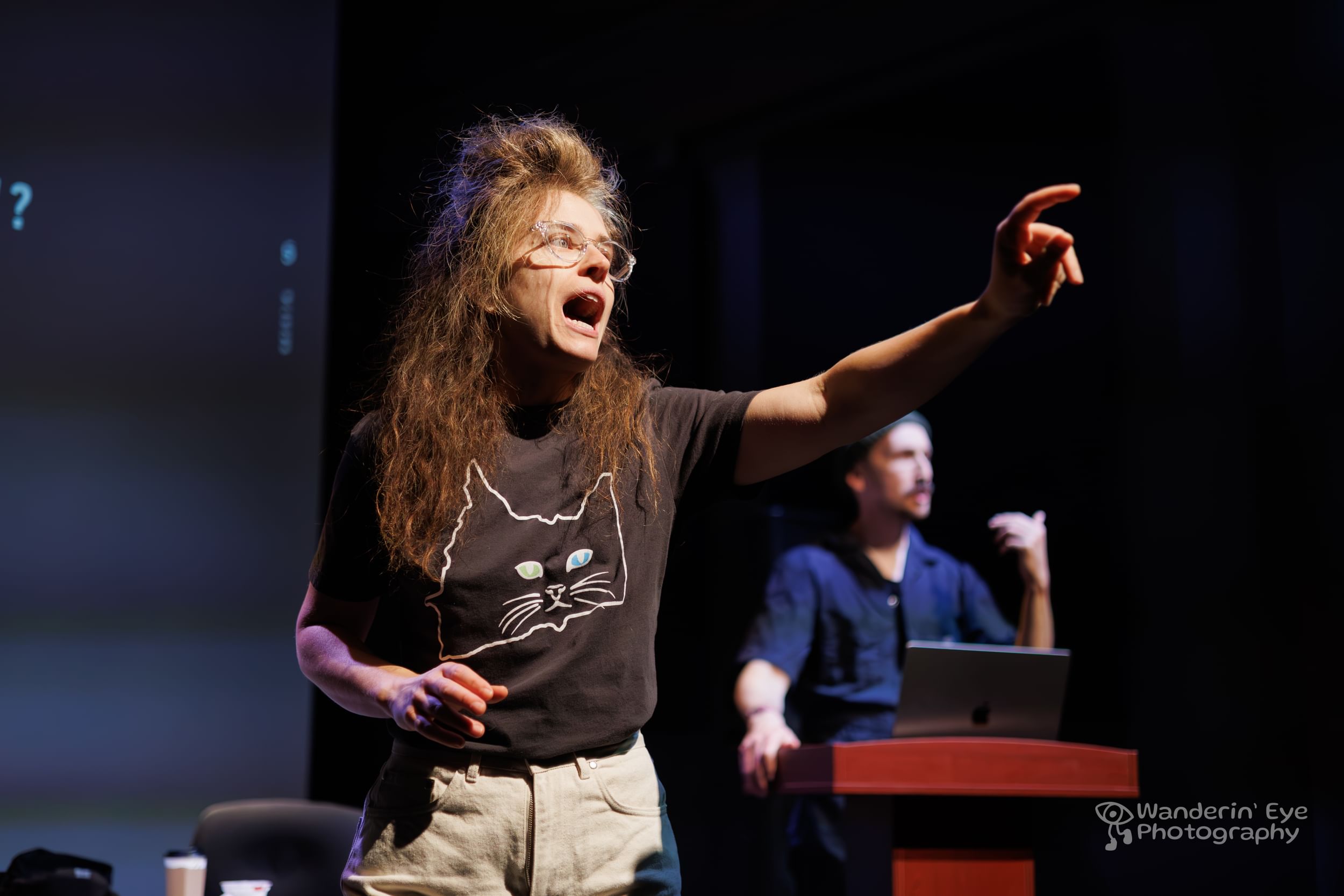 Gold, with long frizzy hair, glasses, and a black tee, gestures dramatically forward, with Braslavsky in the background