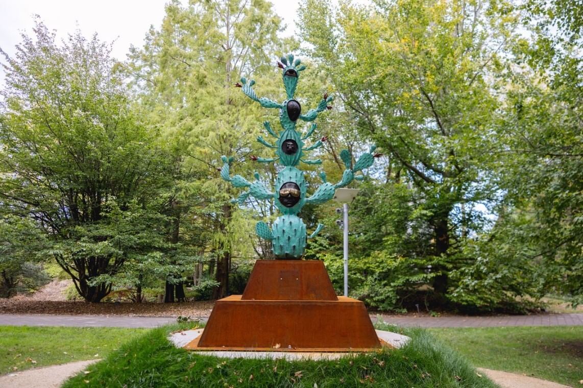 Rusted metal base holds a 9-foot vertical cast bronze blue-green sculpture that melds features of humans and cacti.