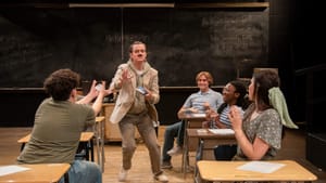 5 students in classroom desks sit around a 6th, who holds a book and wears a fake mustache. There’s a large chalkboard behind