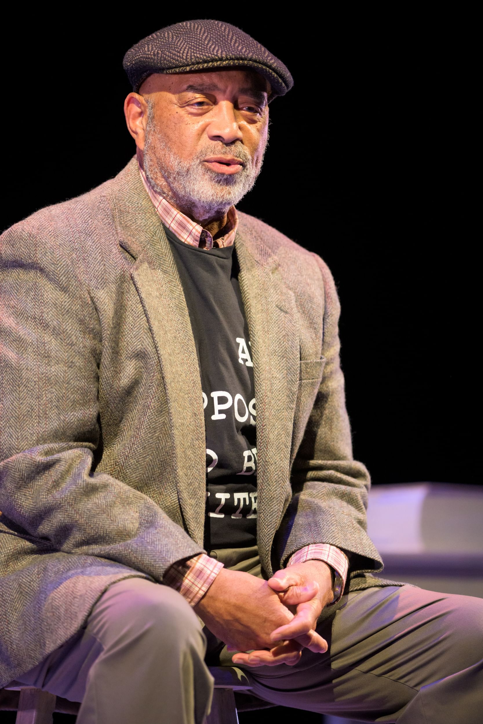 El-Amin, a Black man with a short gray beard, wearing a tweed pageboy hat and blazer, sits on a stool.