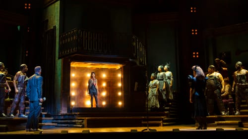 A Black woman actor stands in an elevator-like box onstage, lit with yellow bulbs inside. The ensemble stands watching her.