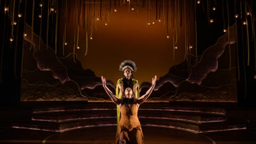 Robinson, in feathered headdress, holds Johnson’s shoulders as she kneels in front with arms up on an earthy, magical stage