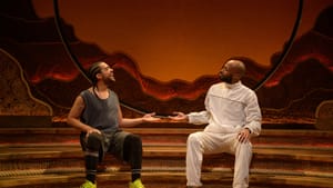 Briggs & Pierce sit on a set of 3 curved steps with subtle orange lighting, in a mirrored pose smiling and extending one hand