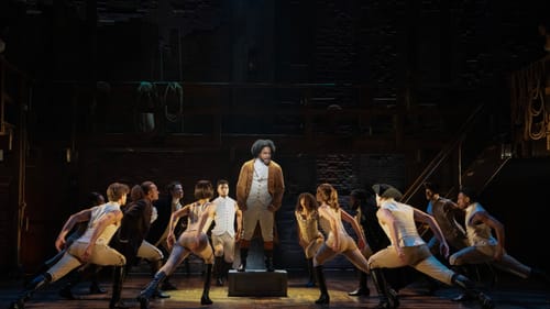 Fauntleroy, in a long brown jacket, sings on a pedestal. The circled ensemble, in white tops & beige leggings, lean to him