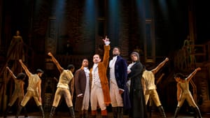 Three male cast members in luxurious costumes stand behind Pierre Jean Gonzalez as Hamilton, victoriously raising one hand