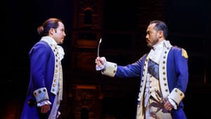 Two male cast members, in splendid blue 18th-century coats, face each other onstage. One has a quill in his outstretched hand