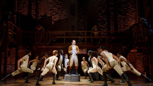 A scene from Hamilton. The actor playing Hamilton stands on a box. The circling ensemble dancers all lean deeply toward him.