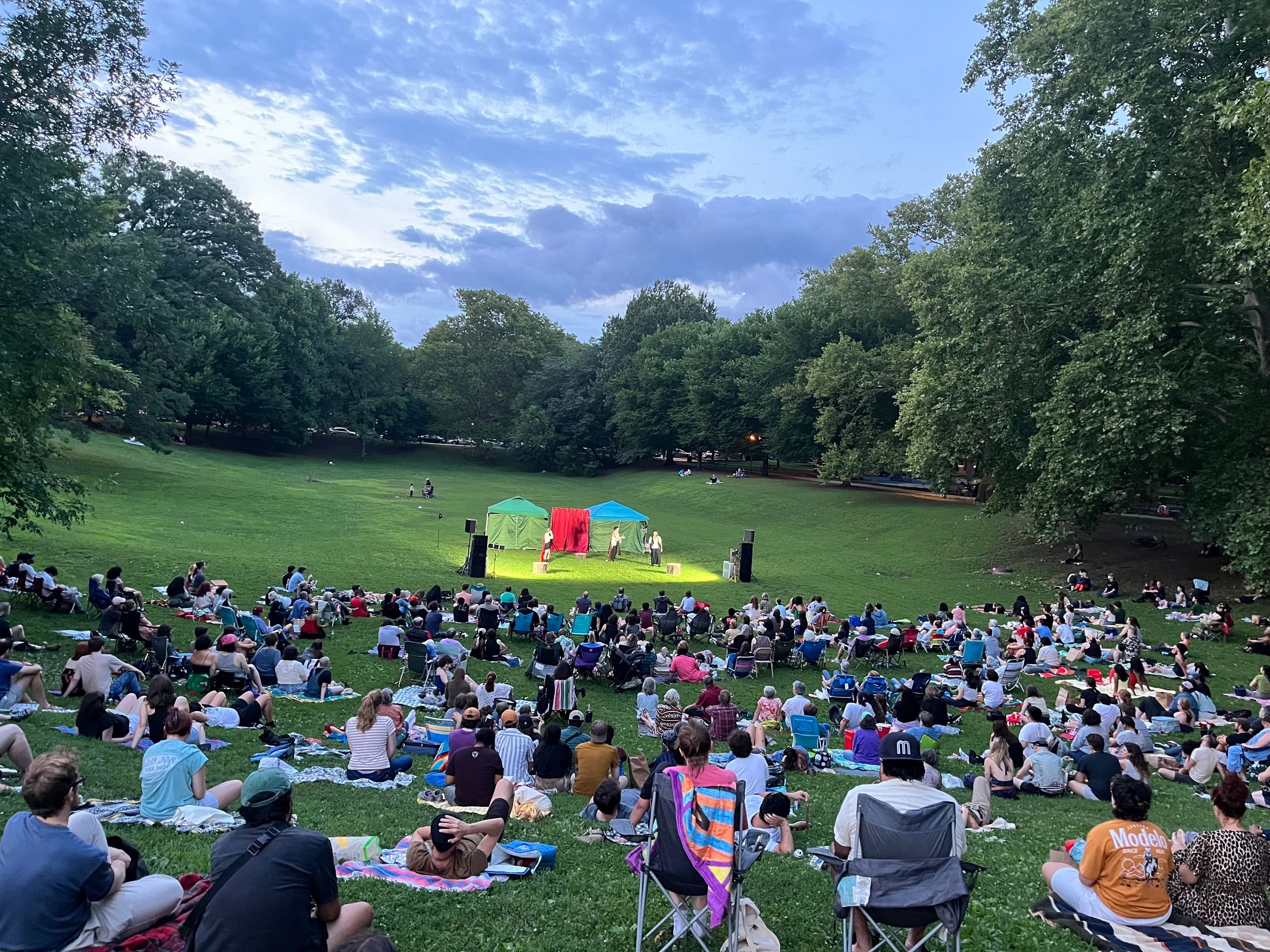 Sunset view of about 350 people sitting in a grassy park with sloping sides, watching the lit-up stage.