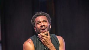 Biko Eisen-Martin, in a sleeveless black costume, kneels on the stage, making a humorous yet anguished face.