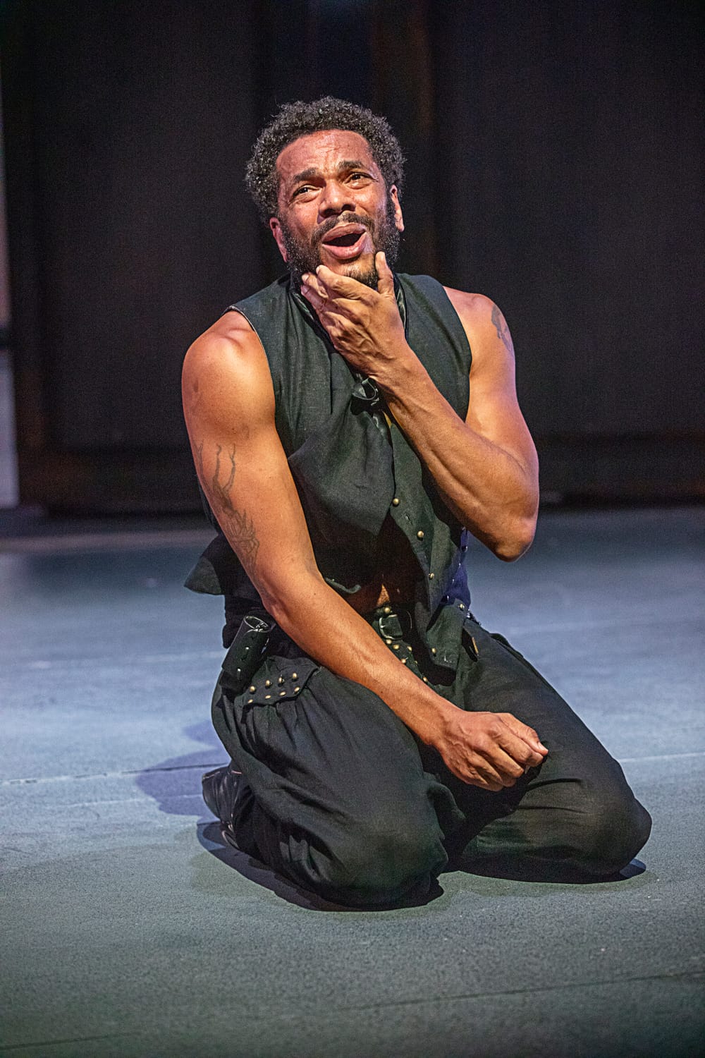 Biko Eisen-Martin, in a sleeveless black costume, kneels on the stage, making a humorous yet anguished face.
