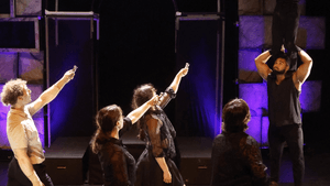 4 actors stand on the ground, 3 of them gesturing to another who perches on high top of two people vertically stacked.