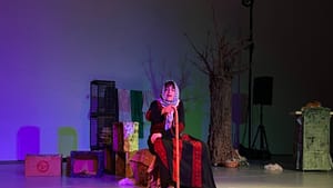 Ali-Ahmad, a Lebanese American woman, sits on a box draped with a rug, holding a cane and wearing a keffiyeh.