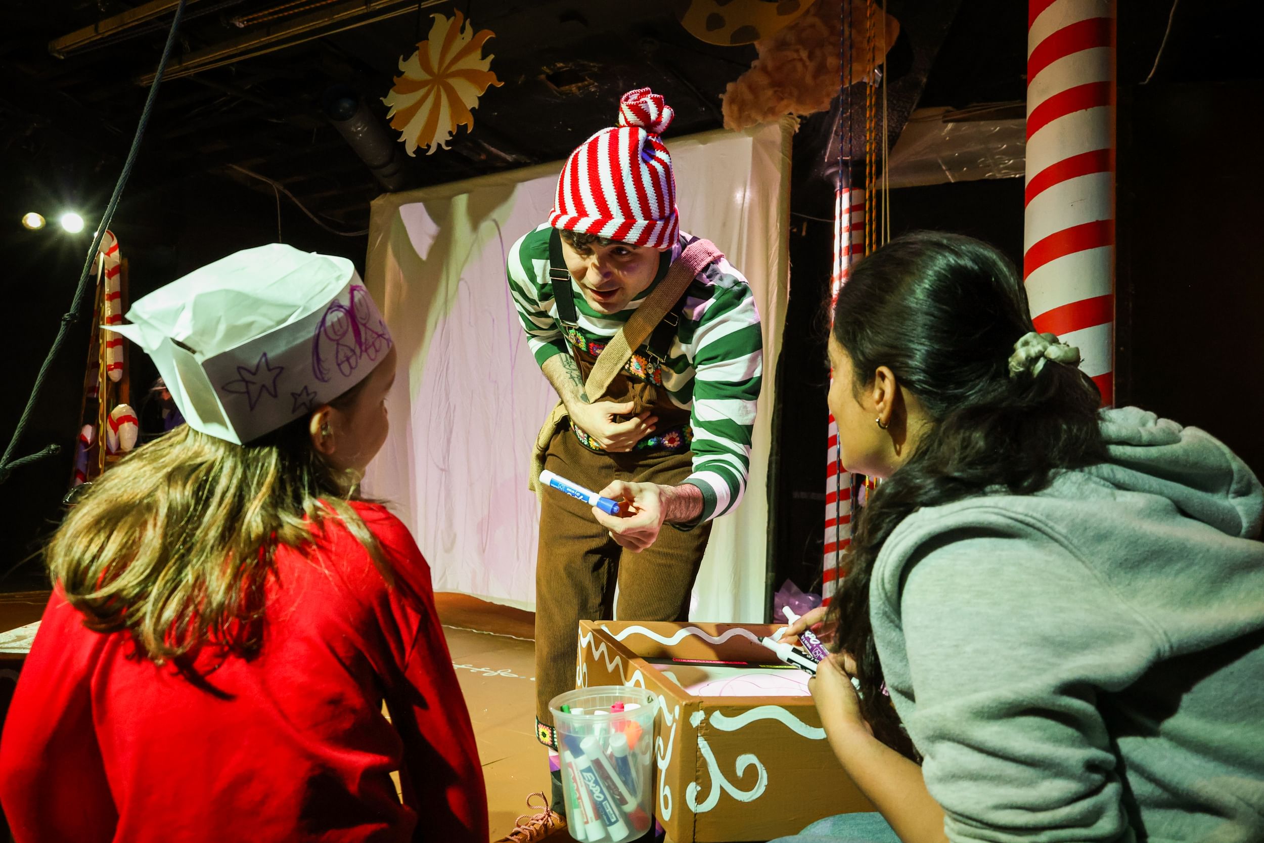 Castagna, in corduroy overalls and red & green stripes, offers a marker to a child wearing a paper hat she has decorated.