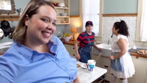 Three people in a contemporary kitchen, one up close looks to camera, the other two as Hansel and Gretel expressing worry