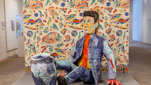 A large colorful ceramic sculpture of a man in a blue suit & orange tie sitting on the ground, his feet tipping an urn over