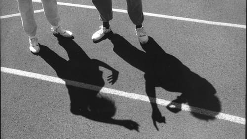 Finkle’s black & white photo, described above. Shadow on pavement of two people in sweats and classic sneakers signing.
