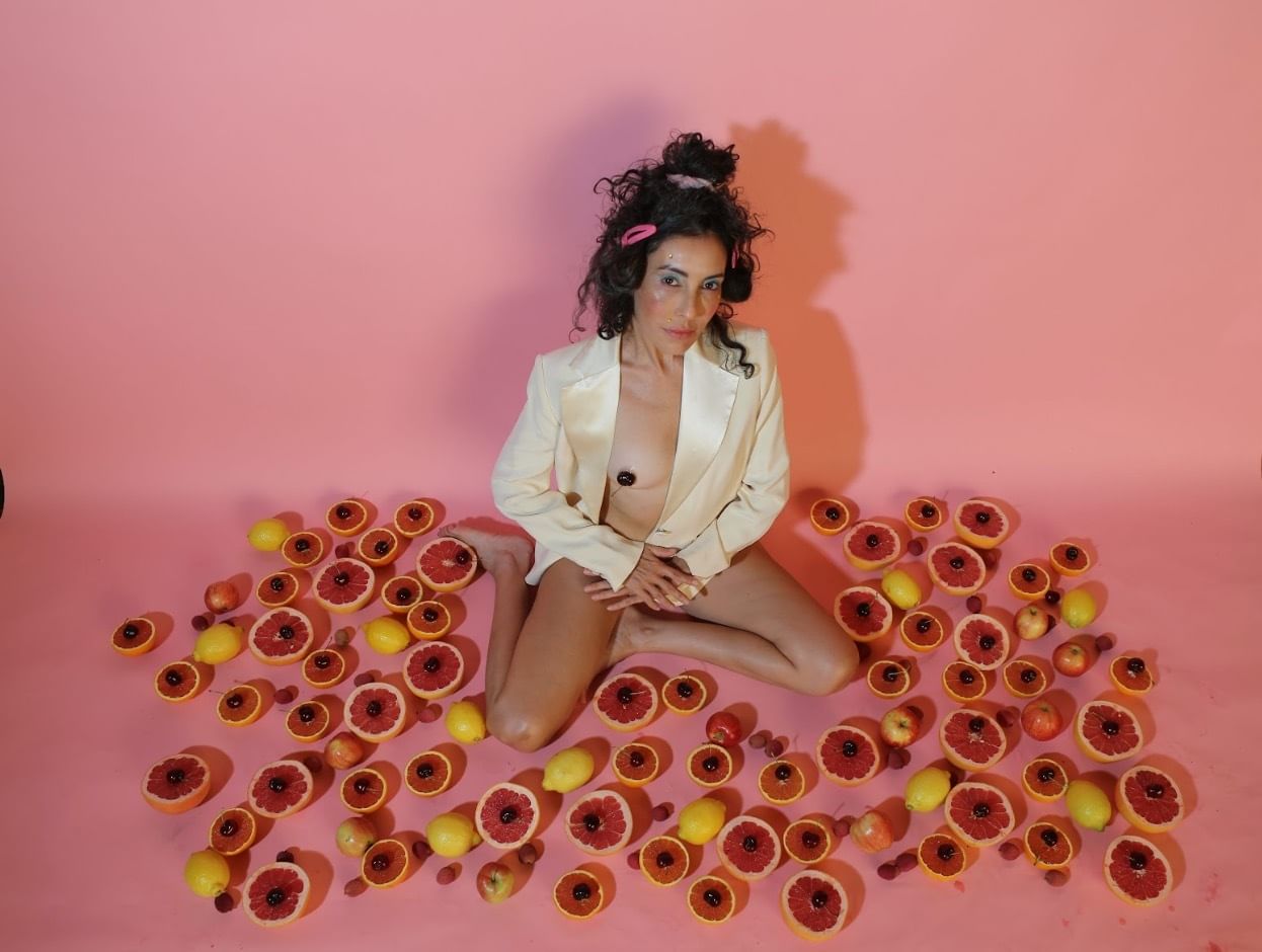 Luardo, hair in a messy updo, wears only a white blazer, sitting on the floor among artfully arranged fruit.