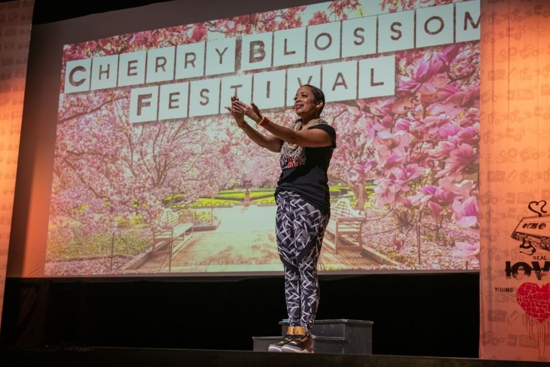 Hernandez, in sneakers, a black tee, and patterned leggings, stands in front of a projection of a Cherry Blossom Festival.